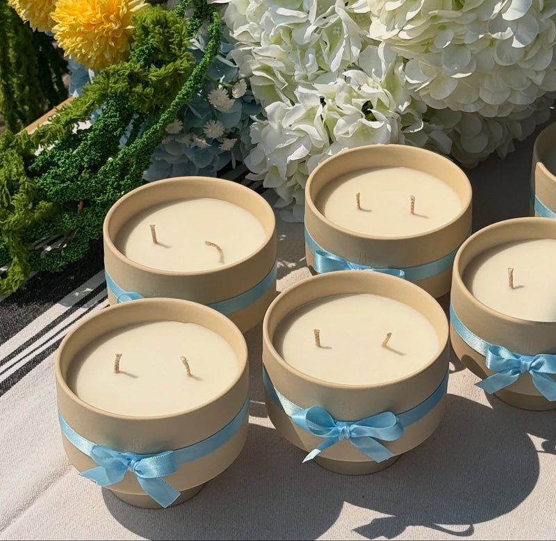 Set of candles in beige containers with blue ribbons, surrounded by flowers on a stone surface.