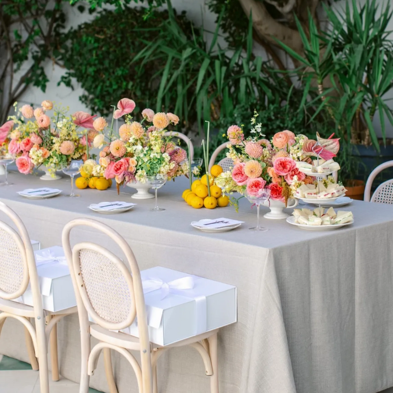 A long outdoor table set with a beige tablecloth, white chairs, floral centerpieces with pink, peach, and yellow flowers, lemons, plates, and a small dessert stand, with leafy greenery in the background.