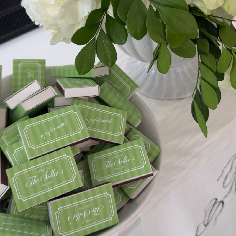 A bowl filled with small green matchboxes printed with the name 'The Seller' and the phrase 'a perfect match,' styled beside a white vase with green leaves and white flowers on a white table.
