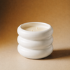 A white ceramic three-tiered candle holder with a double wick candle inside, set against a warm brown gradient background.