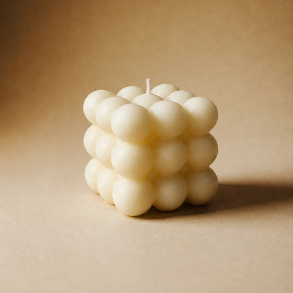 A cream-colored bubble candle shaped like a cube, composed of multiple smooth spherical wax clusters, set against a warm brown textured background.